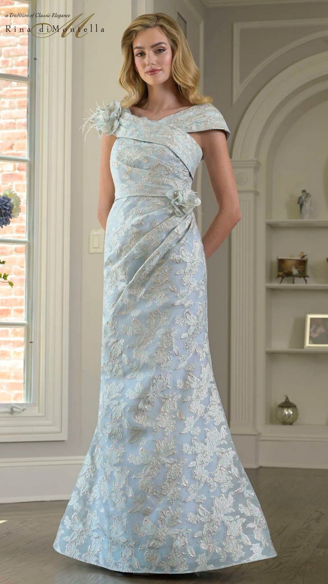 A woman stands indoors wearing the Rina Di Montella RD3428 long formal dress in blue, featuring silver jacquard patterns and floral embellishments on the shoulder and waist.